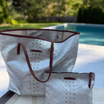 White tote bag with brown accents and a matching pouch on a table by a pool.