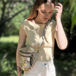 Woman wearing a beige top and white pants with a gold fanny pack in a garden setting