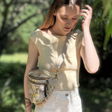 Woman wearing a beige top and white pants with a gold fanny pack in a garden setting