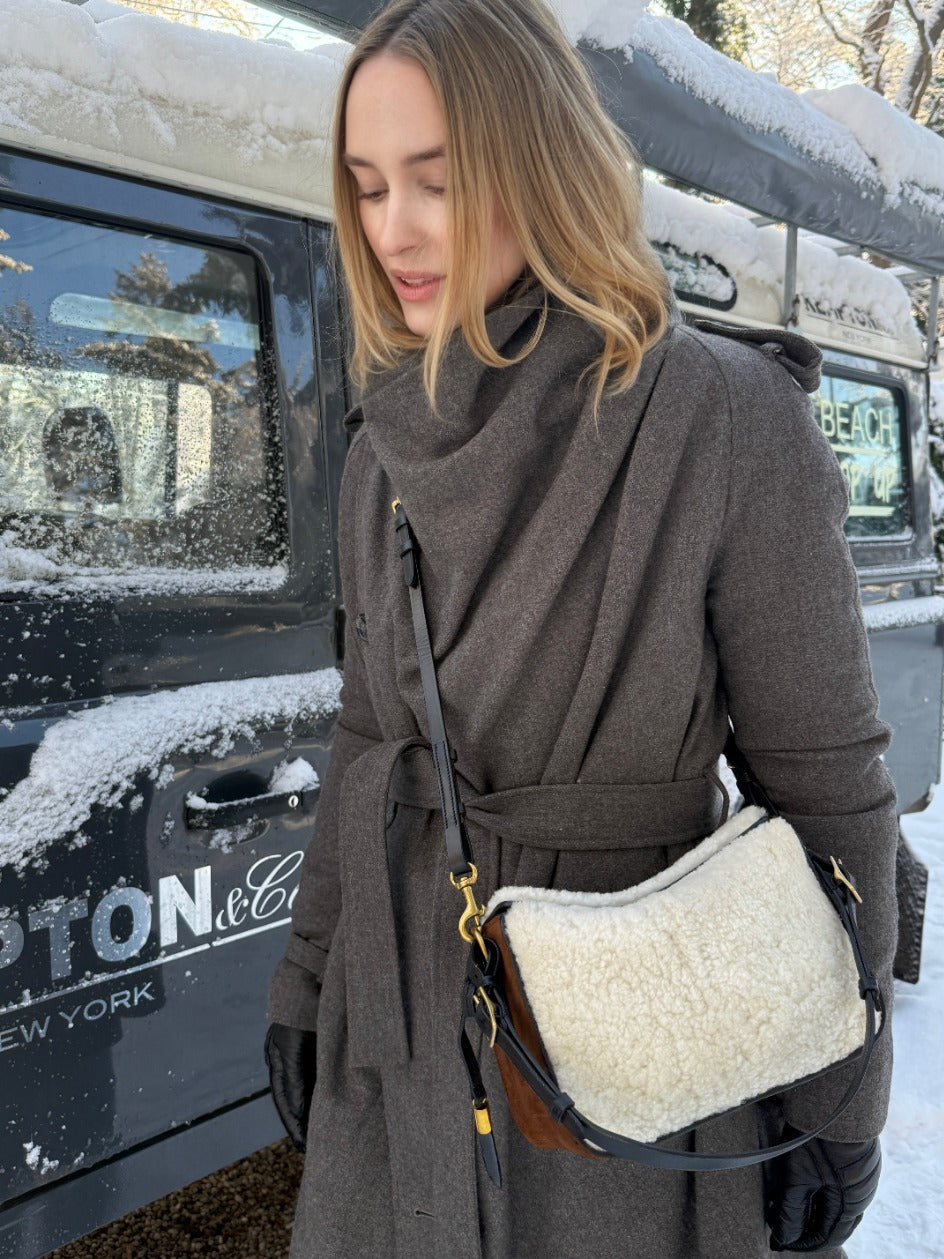 A woman wearing a dark brown belted coat and black gloves carries a cream sheepskin shoulder bag with brown leather trim and gold hardware, standing outdoors next to a snow-dusted black vehicle.
