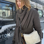 A woman wearing a dark brown belted coat and black gloves carries a cream sheepskin shoulder bag with brown leather trim and gold hardware, standing outdoors next to a snow-dusted black vehicle.